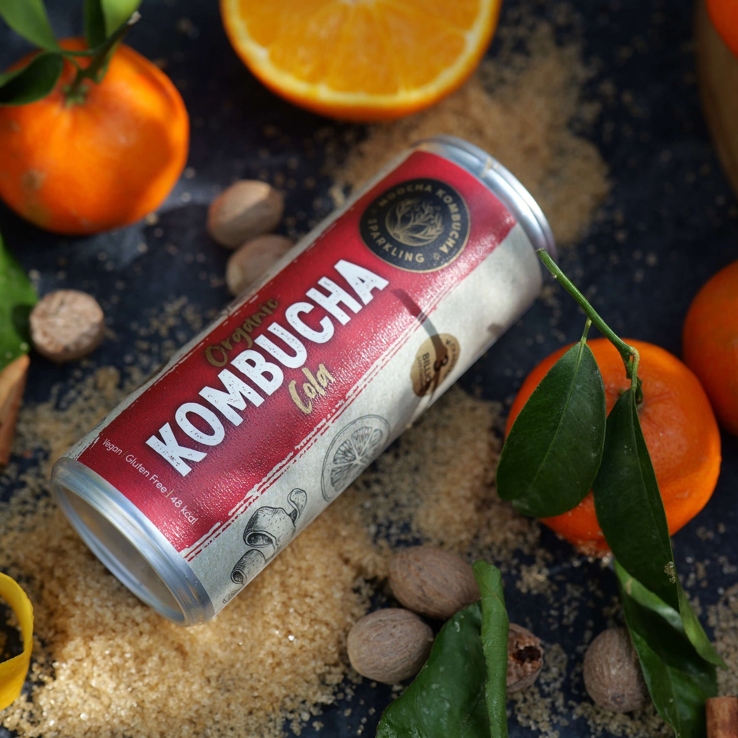 a can of cola kombuhca on a black slate on a bed of brown sugar, surrounded with slices of oranges and balls of nutmeg spice