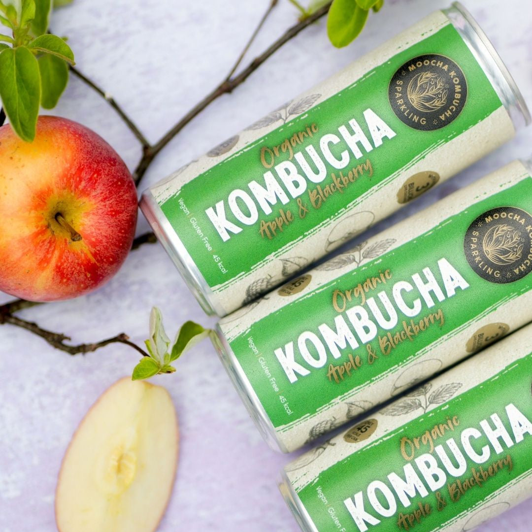Three cans of Organic Kombucha Apple & Blackberry flavor with apples on a light background