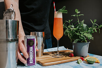 mixed berry moocha kombucha, a large stem glass with a refreshing kombucha drink and red and white straw with a guarnis of mint om the top, while a man holds a can of moocha kombucha 